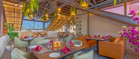A restaurant with high ceilings, hanging plants, and a view of the city. Tables are set with red napkins and glassware.