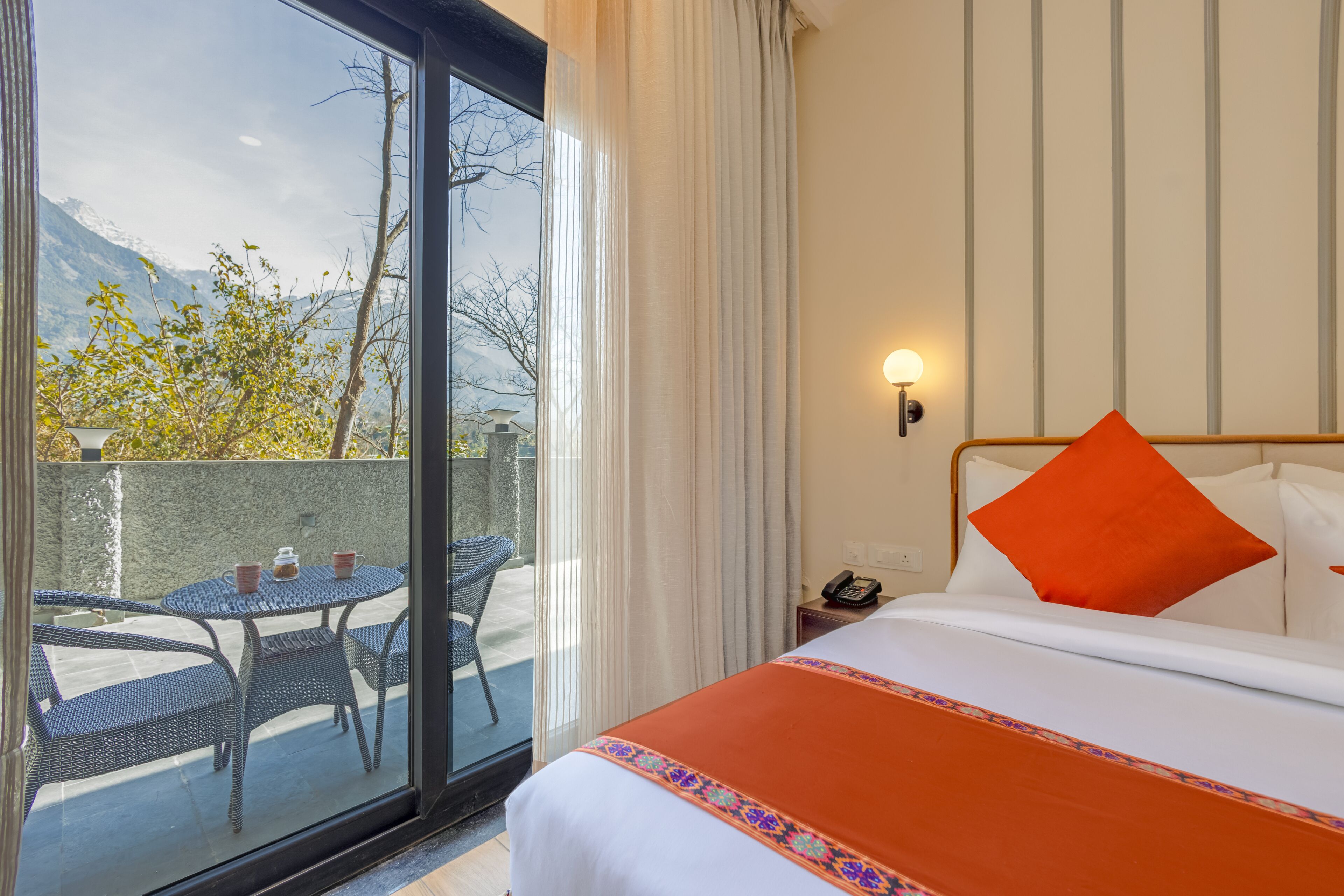 Une chambre d’hôtel comprenant un lit, une table de chevet, un téléphone, une table avec des chaises et une vue sur les montagnes, visible à travers une porte vitrée.