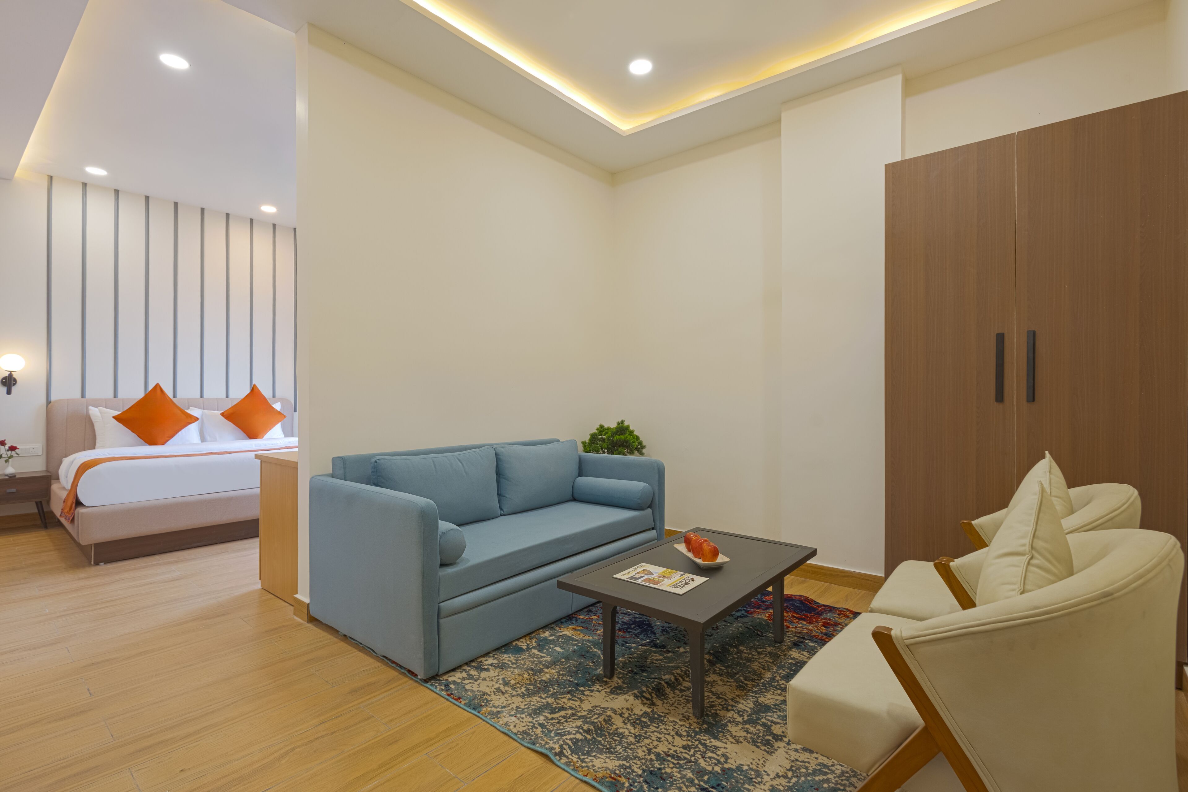 A modern hotel room with a bed, a blue sofa, a white armchair, a coffee table, and a wardrobe.