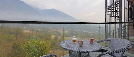 En balkon med fletningsmøbler og gelænder i glas, med udsigt over et bjerglandskab.