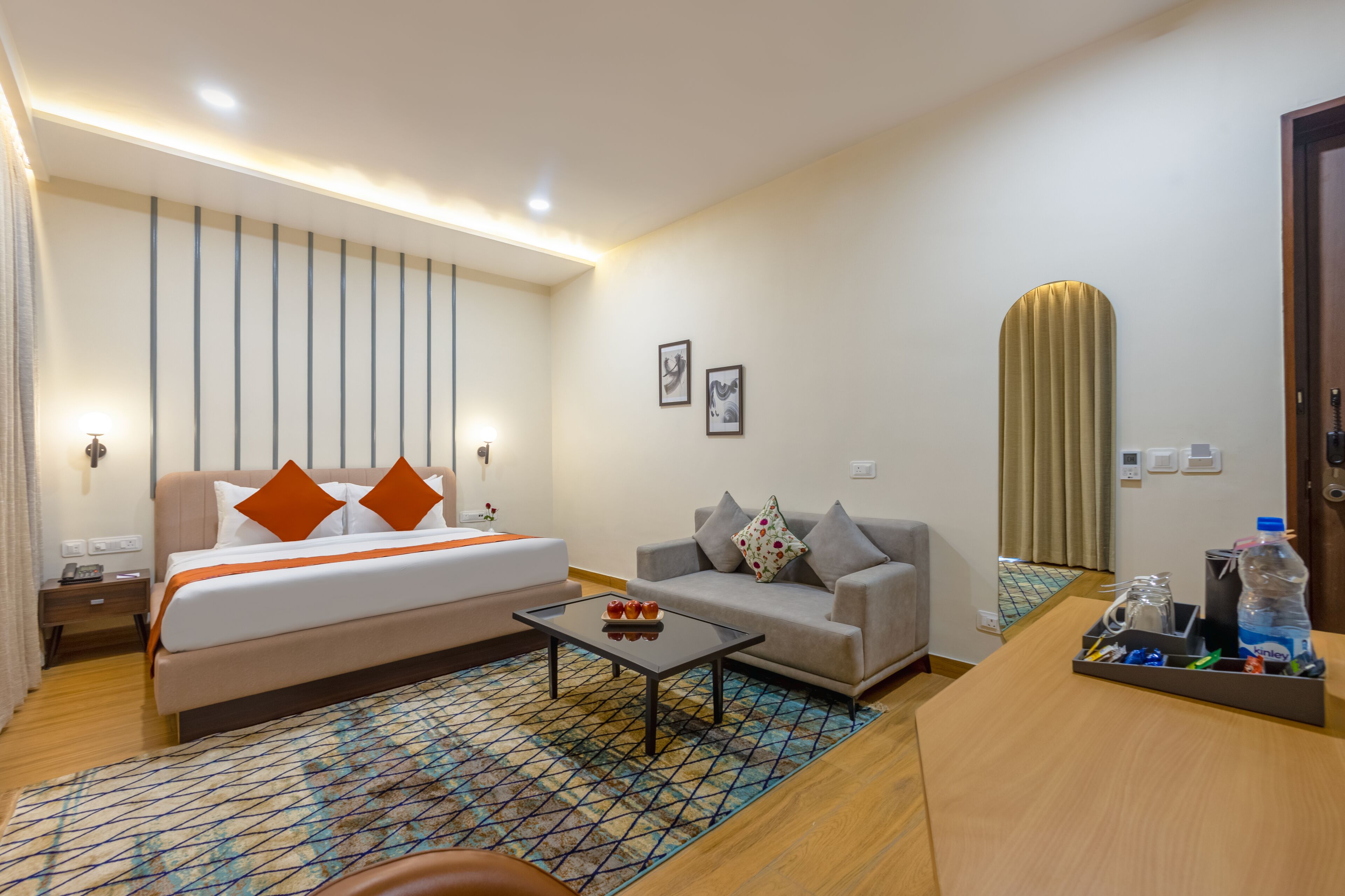Habitación de hotel con cama, sofá, mesita de centro y escritorio con una botella de agua y vasos.