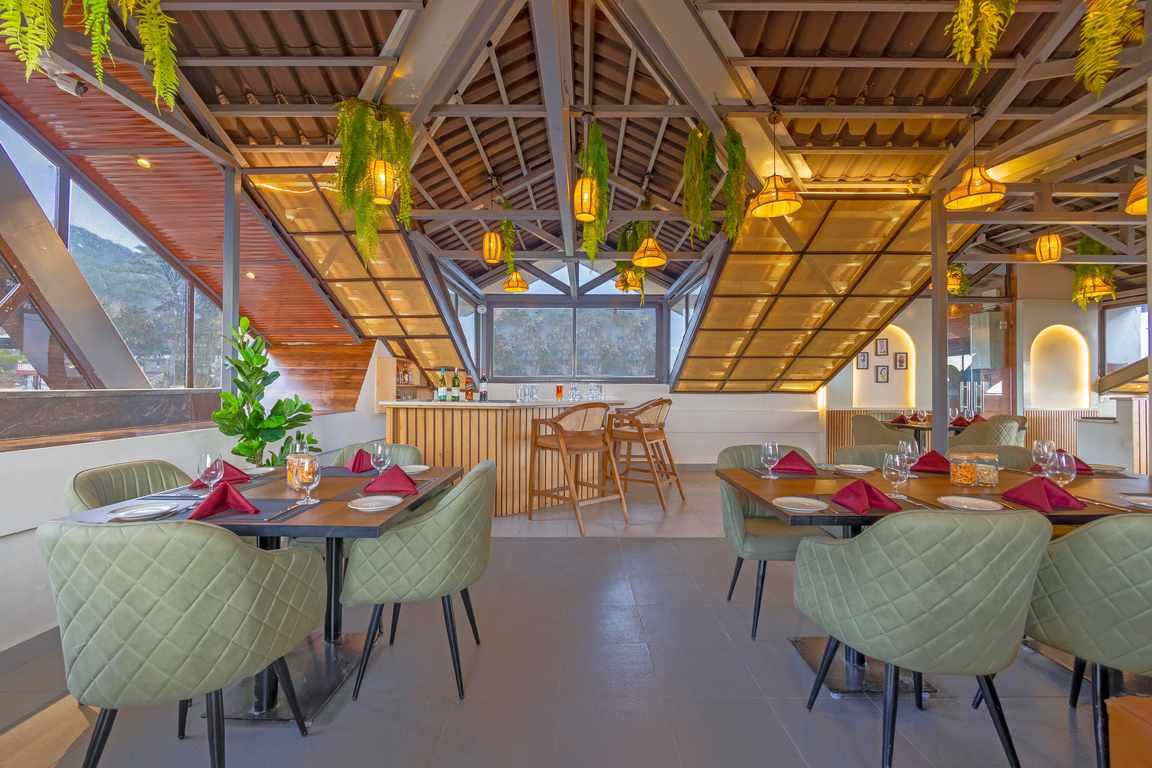 A restaurant with a high ceiling, hanging plants, and a view of the mountains. Tables are set with red napkins and glassware.