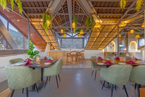 Een restaurant met een hoge plafond, hangende planten en uitzicht op de bergen. Tafels zijn gedekt met rode servetten en glazen.