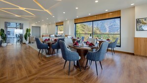 A modern dining area with a large table, blue chairs, and a view of mountains through large windows. - Sterling Nirmaya Dharamshala (Dharamshala)