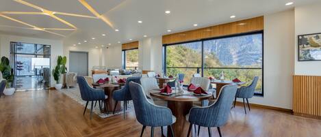 A modern dining area with a large table, blue chairs, and a view of mountains through large windows.