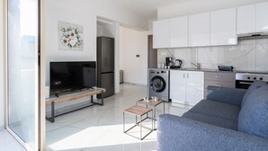 Una sala moderna con un sofá gris, un televisor de pantalla plana y una mesa de centro. Se ve un área de cocina con armarios y electrodomésticos blancos al fondo.
