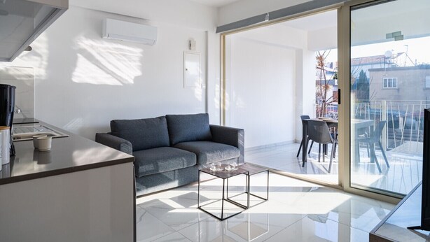 Un apartamento moderno con un sofá gris, una pequeña mesa de centro, un balcón con vistas a la ciudad y una zona de cocina.
