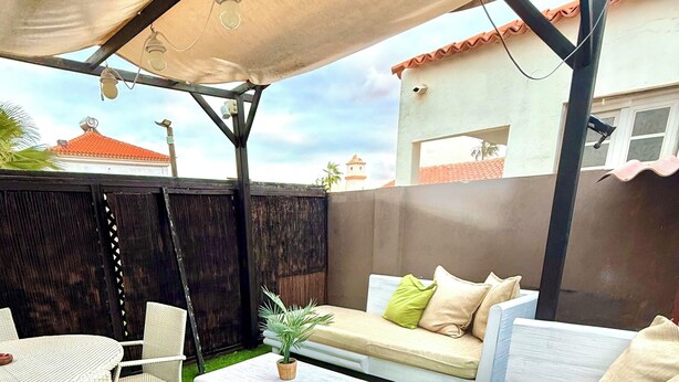 Una zona patio con tavolo e sedie bianche, un divano beige con cuscini, un piccolo tavolino e una pianta in vaso.