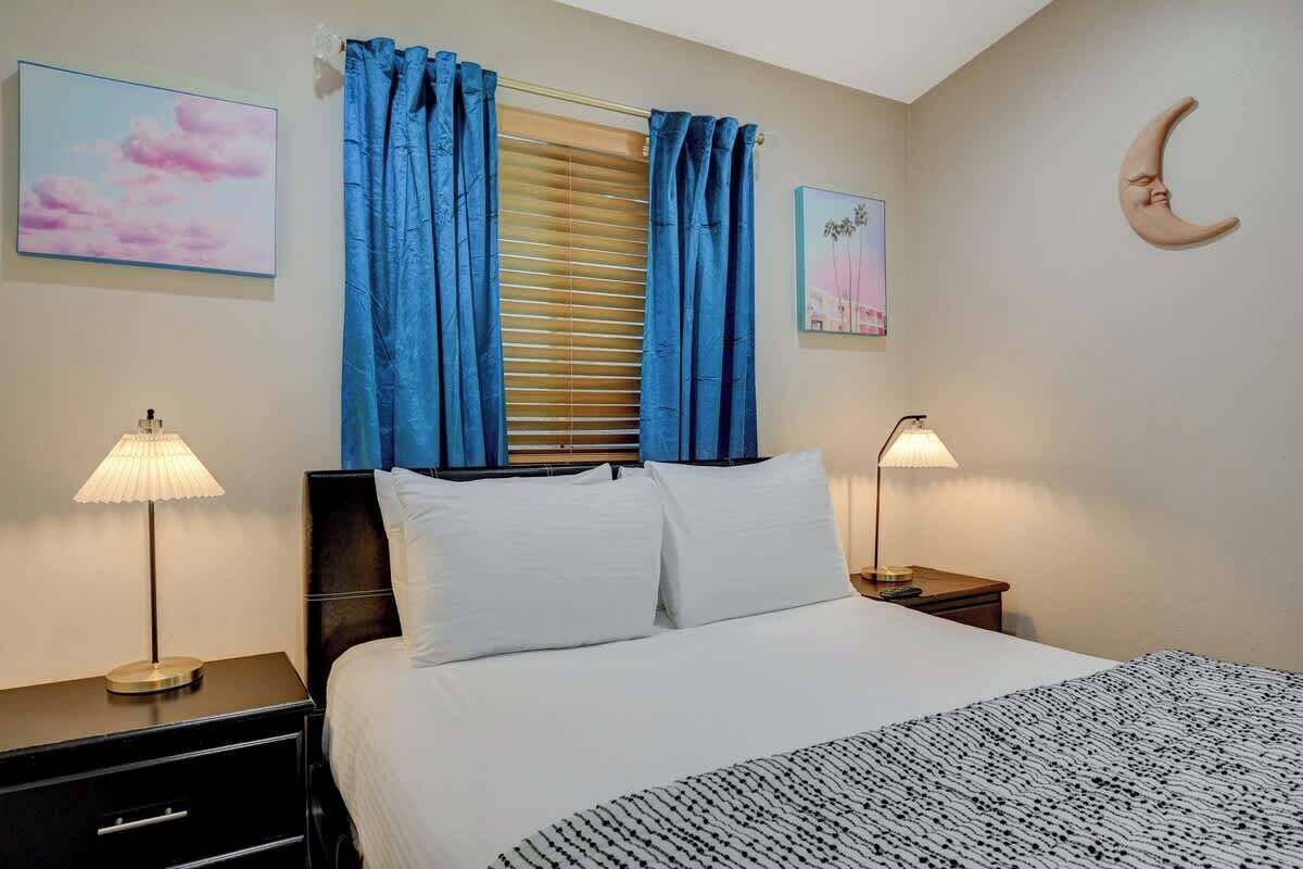 Een slaapkamer met een bed, twee nachtkastjes, een raam met blauwe gordijnen en wanddecoratie.
