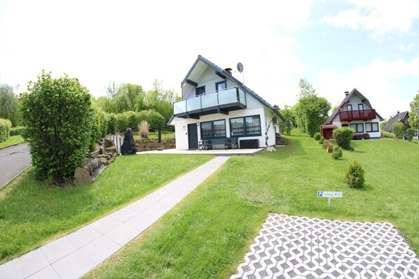 A modern two-story house with a balcony, a paved driveway, and a small garden area.