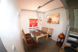 A dining area with a wooden table, four chairs, a hanging light fixture, and a wall-mounted picture.