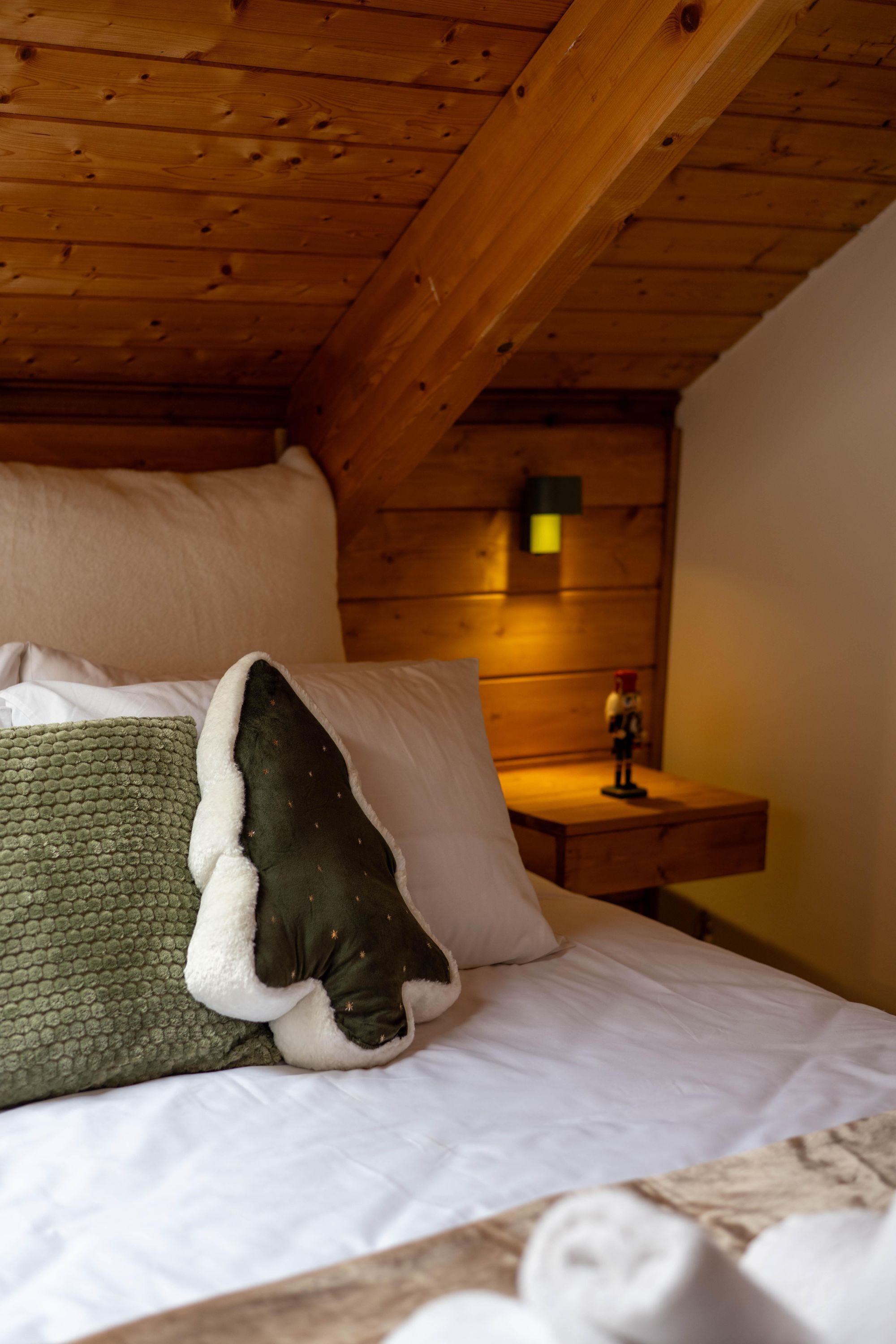 Un plafond en bois, un lit avec des oreillers blancs et verts, une table de chevet avec une lampe et une petite figurine.
