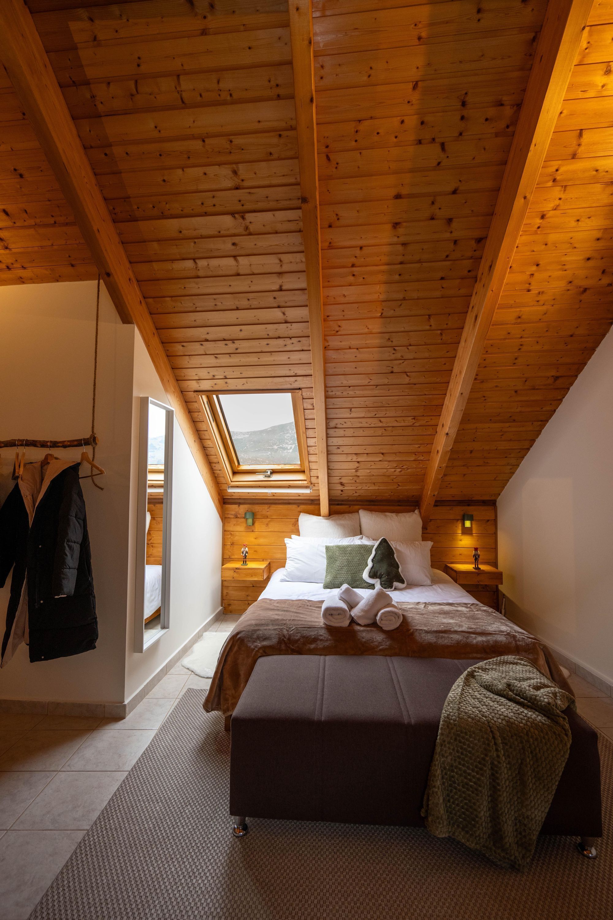 Une chambre confortable avec un plafond en pente, un lit avec des oreillers et une couverture, une table de chevet avec une lampe et un porte-manteau avec une veste.
