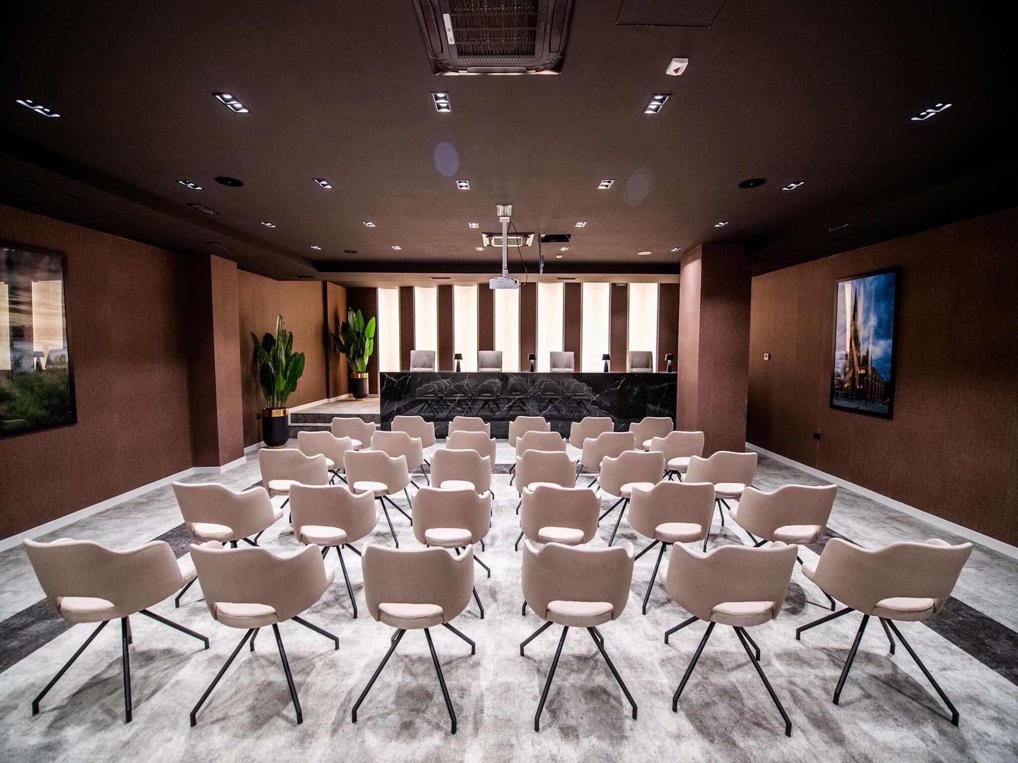 A conference room with rows of chairs facing a reception desk, large windows, and a ceiling with recessed lighting.