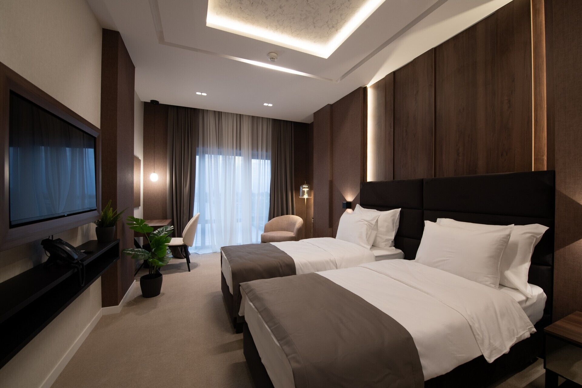 Ein modernes Hotelzimmer mit zwei Betten, einem Fernseher, einem Schreibtisch und einem großen Fenster mit Vorhängen.