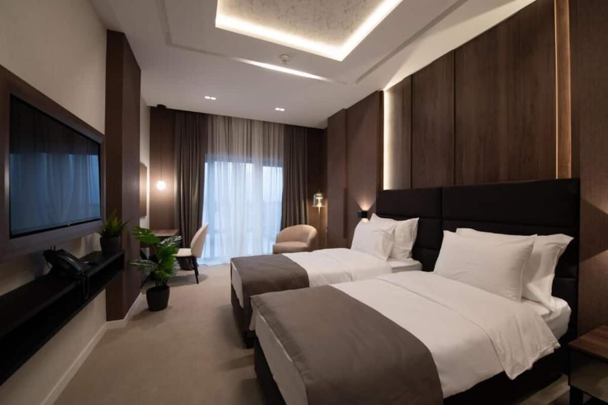 A modern hotel room with two beds, a flat-screen TV, a desk with a telephone, and a seating area with a chair and curtains.