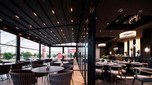Ein modernes Restaurant mit runden und rechteckigen Tischen, schwarzen und grauen Stühlen sowie einem großen Fenster mit Blick auf die Stadt.