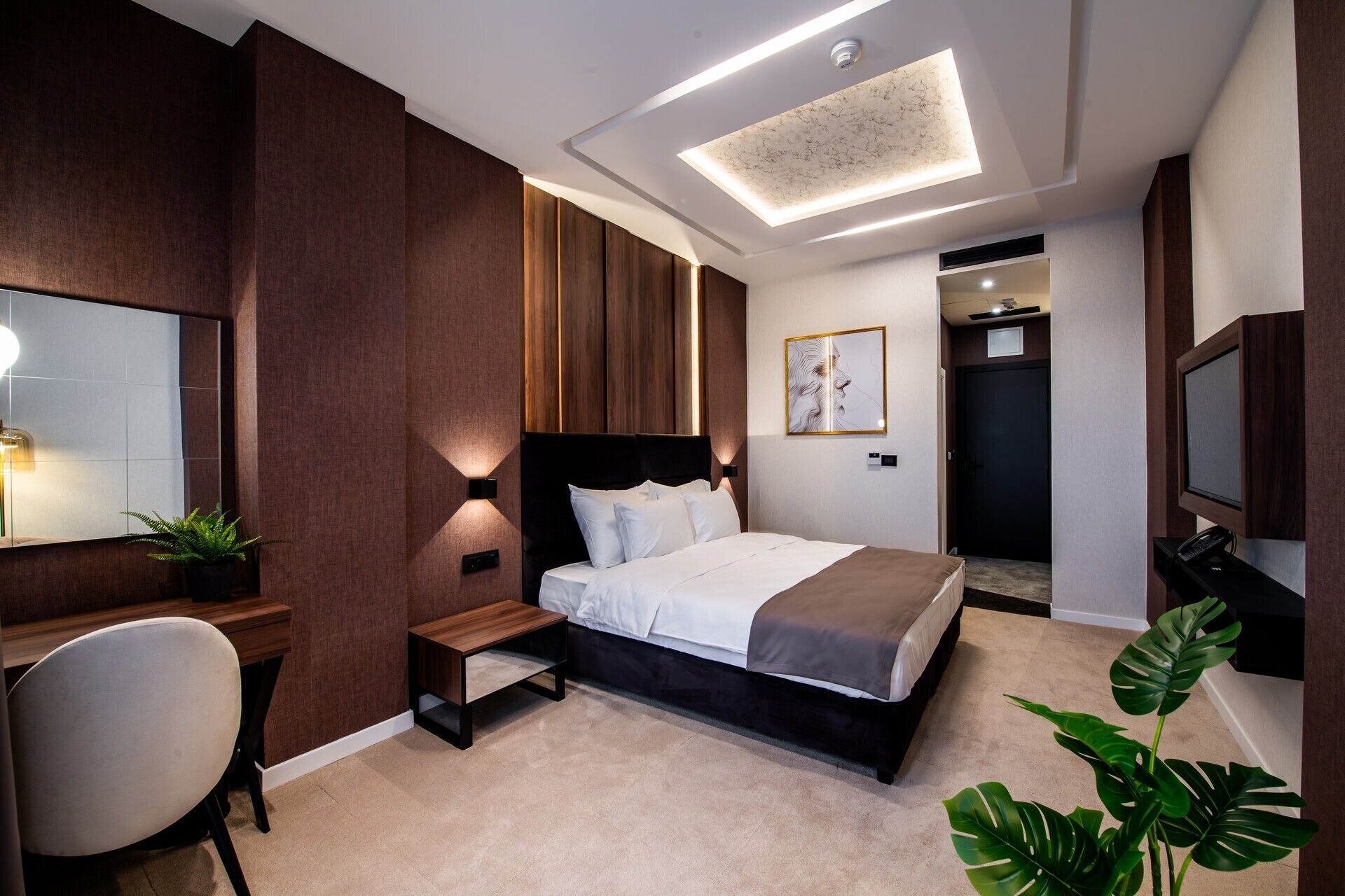A modern hotel room with a large bed, a desk with a chair, a small table, and a large mirror. There is a ceiling light and a plant on the desk.