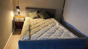 Een net opgemaakt bed met een bloemenprintdeken, een klein nachtkastje met een lamp, en een donker hoofdbord.