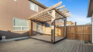 Una terraza de madera con una estructura de pérgola, una valla de madera y una casa con revestimiento horizontal.