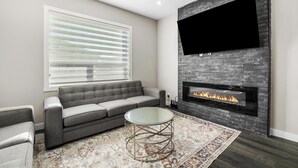 Una sala de estar moderna con un sofá gris, una mesa de centro de vidrio, una alfombra estampada, una chimenea y un televisor montado.