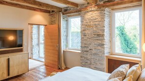 Ein Schlafzimmer mit Steinwänden, Holzbalken, einem Bett, einem Fernseher und einem Fenster mit Blick auf Grünflächen.