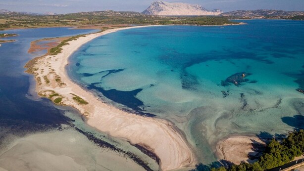 Luchtbeeld van een kustgebied met een zandstrand, helderblauw water en een berg op de achtergrond.