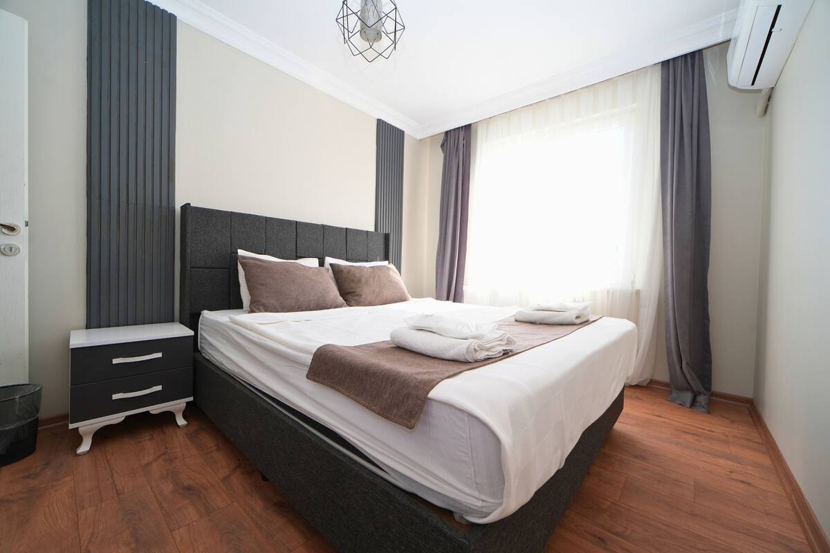 A modern bedroom with a large bed, a bedside table, a wardrobe, and a window with curtains.