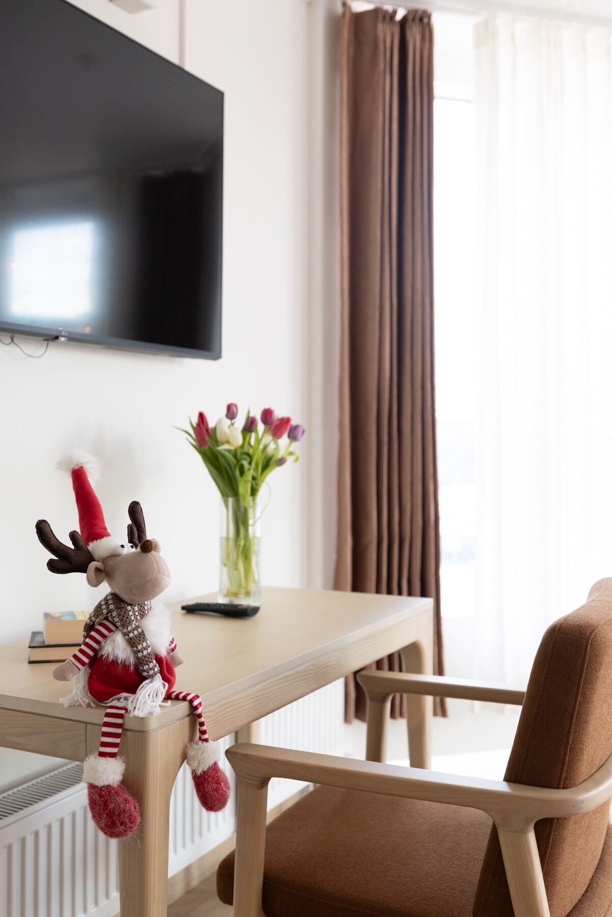 A hotel room with a wooden table, a plush reindeer toy, a vase with flowers, a brown chair, and a flat-screen TV mounted on the wall.