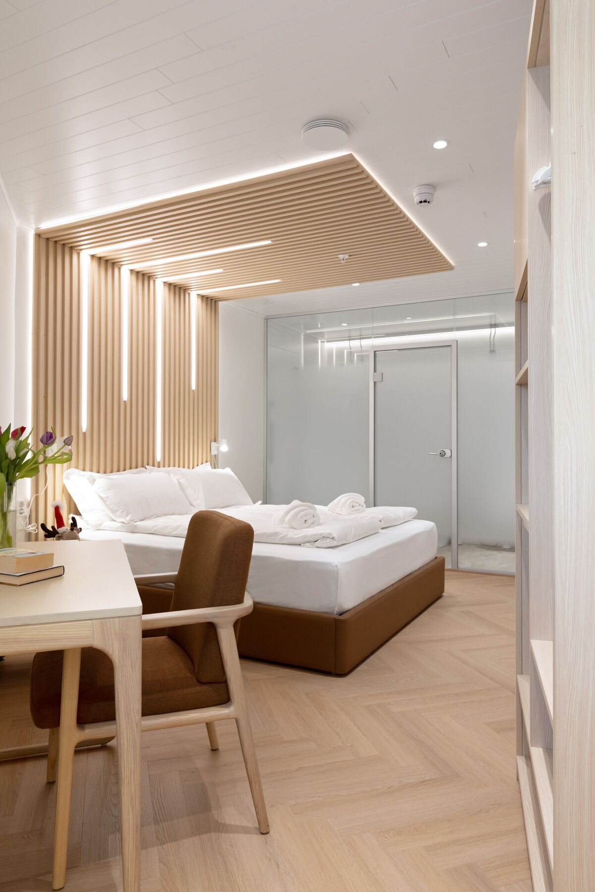 Ein modernes Hotelzimmer mit Holzbalkendecke, einem Bett mit weißen Leinen, einem Schreibtisch mit Stuhl und einer Glastür zum Badezimmer.