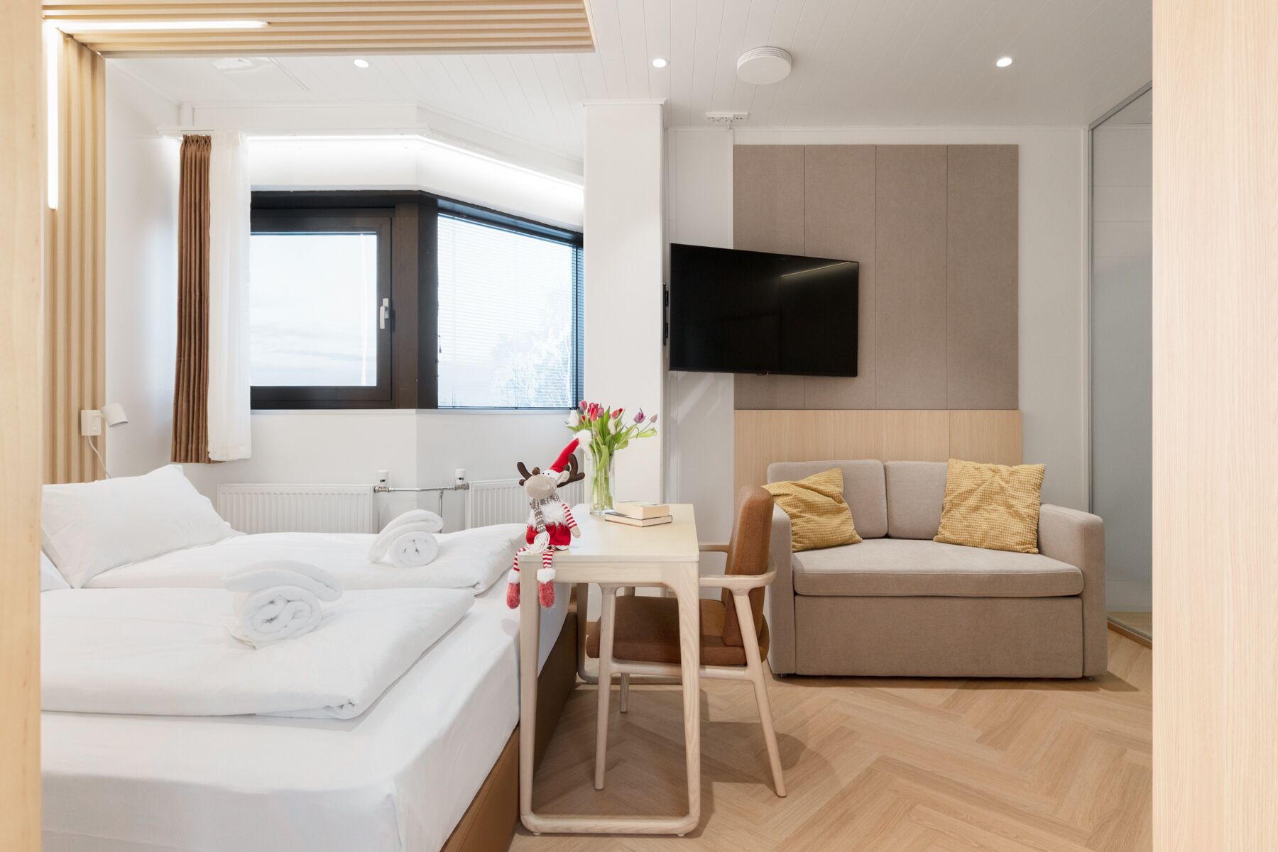 A modern hotel room with a bed, a small table with a vase of flowers, a sofa, and a TV mounted on the wall.