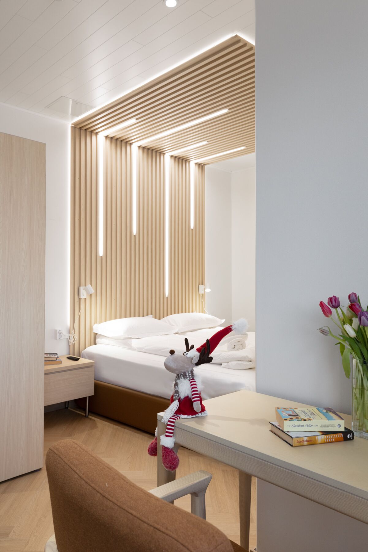 A modern hotel room with a wooden slatted ceiling, a bed with white linens, a plush toy on the bed, a desk with books, and a vase with flowers.