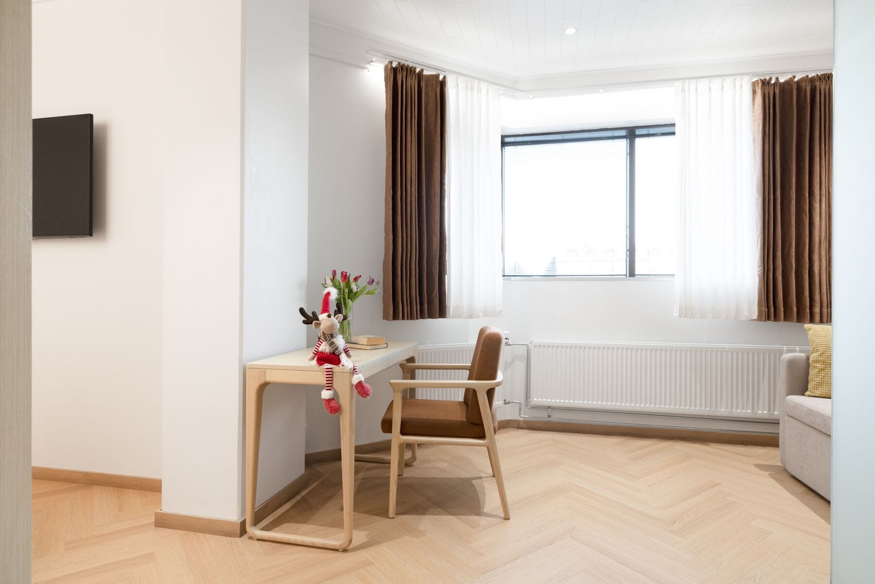 A modern room with a wooden floor, a desk with a chair, a radiator, and a window with curtains.