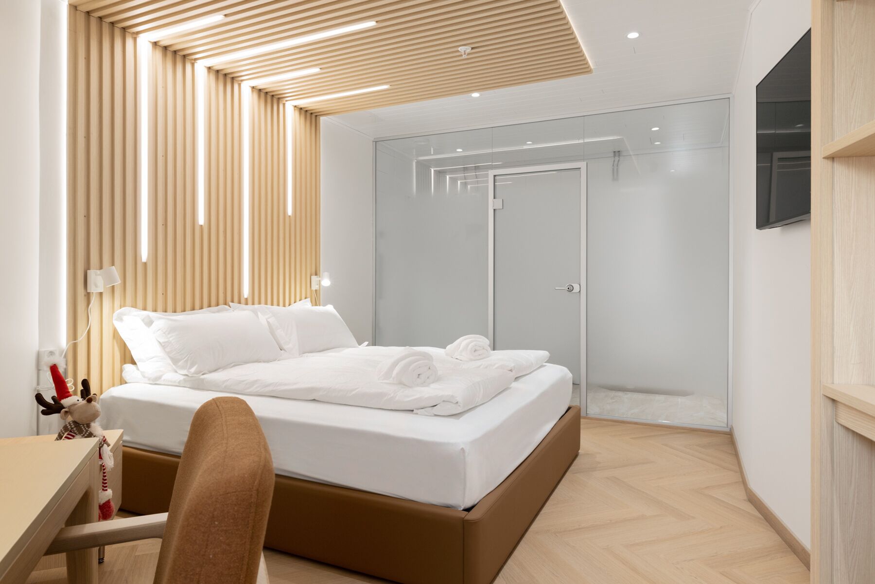 A modern hotel room with a wooden slatted ceiling, a bed with white linens, a glass shower enclosure, and a mounted TV.