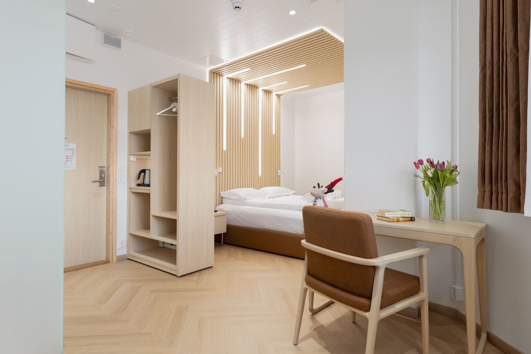 A modern hotel room with a bed, a desk, a chair, a wardrobe, and a window with curtains.