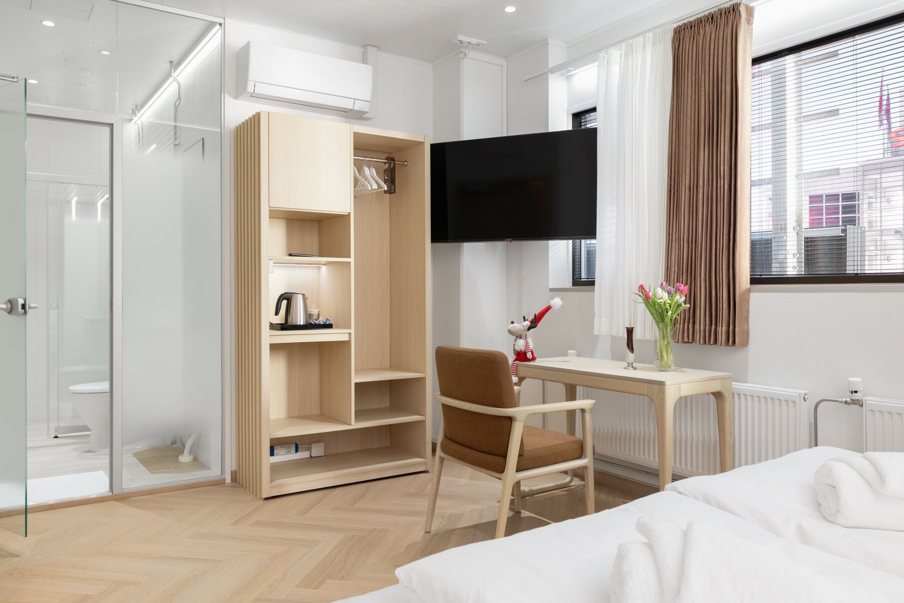 A modern hotel room with a bed, a desk with a chair, a wardrobe, a flat-screen TV, and a window with curtains.