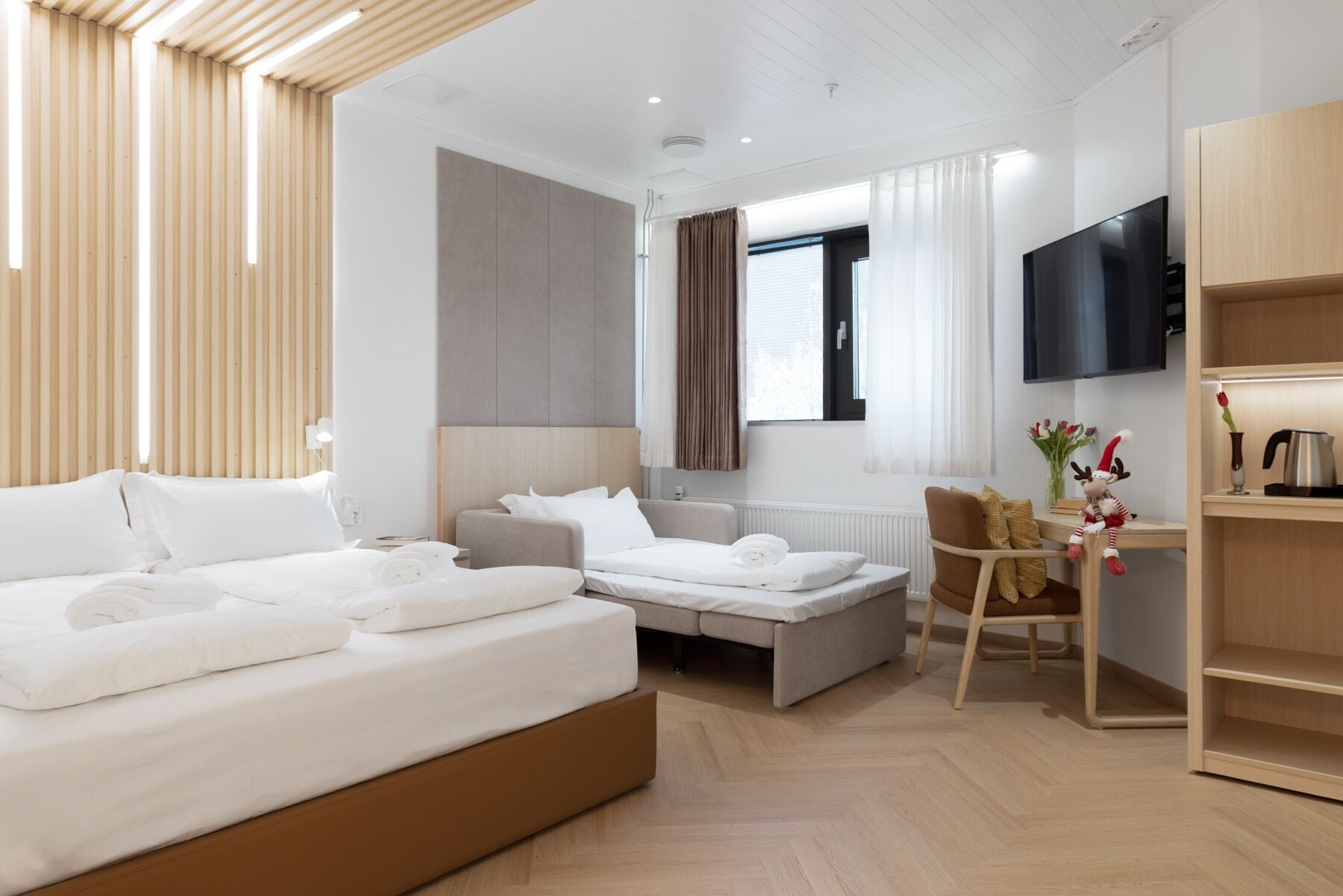 Habitación de hotel moderna con una cama grande, un sofá, un televisor y una pequeña mesa con un objeto decorativo.