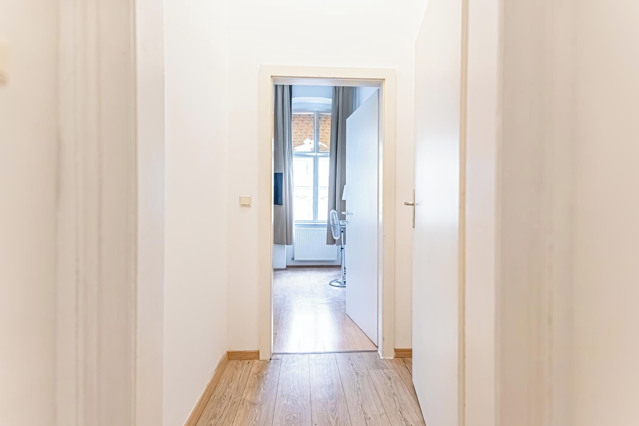 A narrow hallway with a wooden floor leading to a room with a radiator and a window.