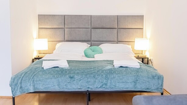 Ein ordentlich bezogenes Bett mit grauem Kopfteil, weißen Kissen und einem grünen Zierkissen. Auf jeder Seite des Bettes stehen zwei Nachttischlampen.