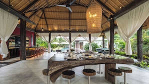 Een overdekt terras met een eethoek, een bar, houten krukken en een zwembad op de achtergrond.