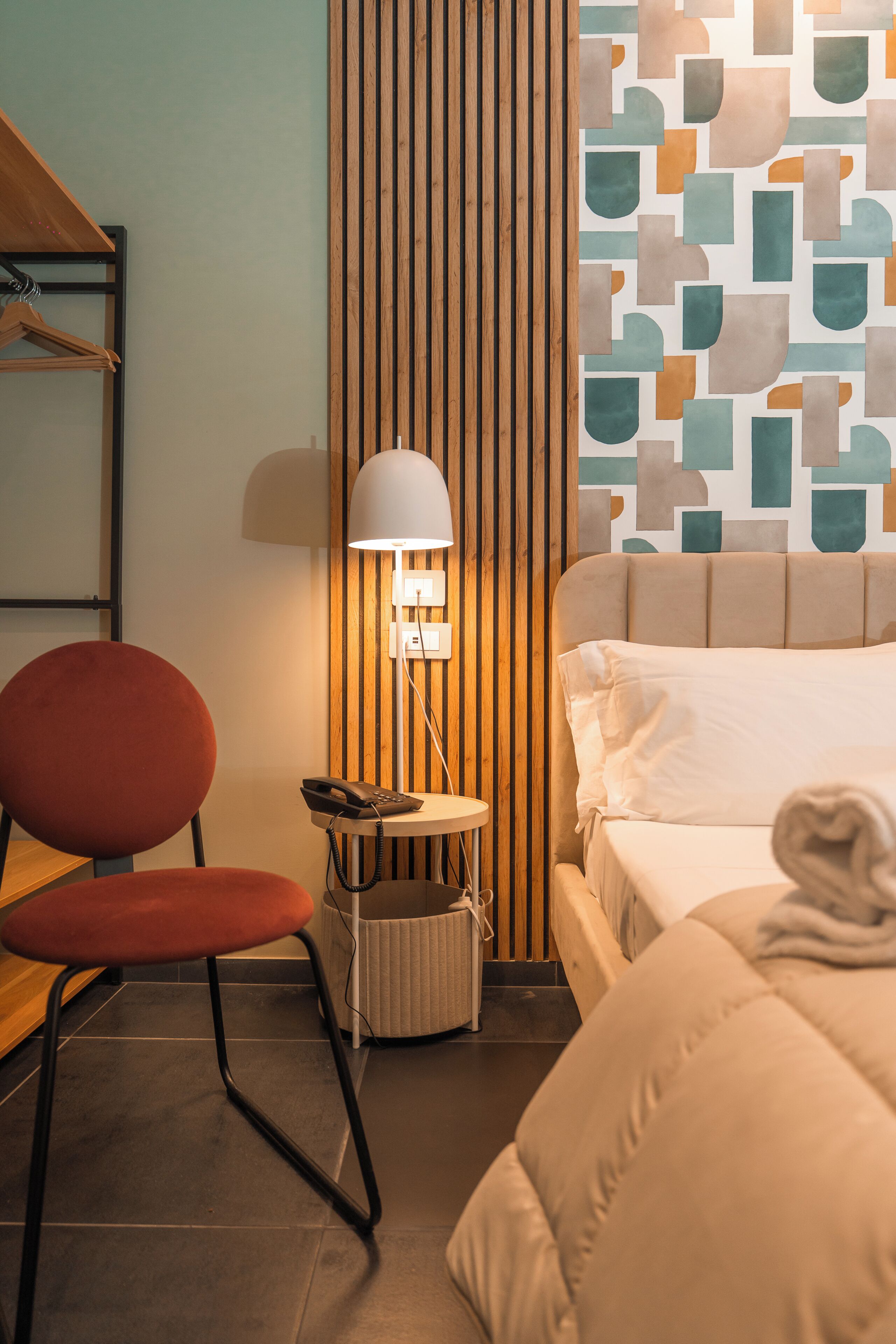 Habitación de hotel con cama, escritorio, silla, lámpara y pared con diseño geométrico.