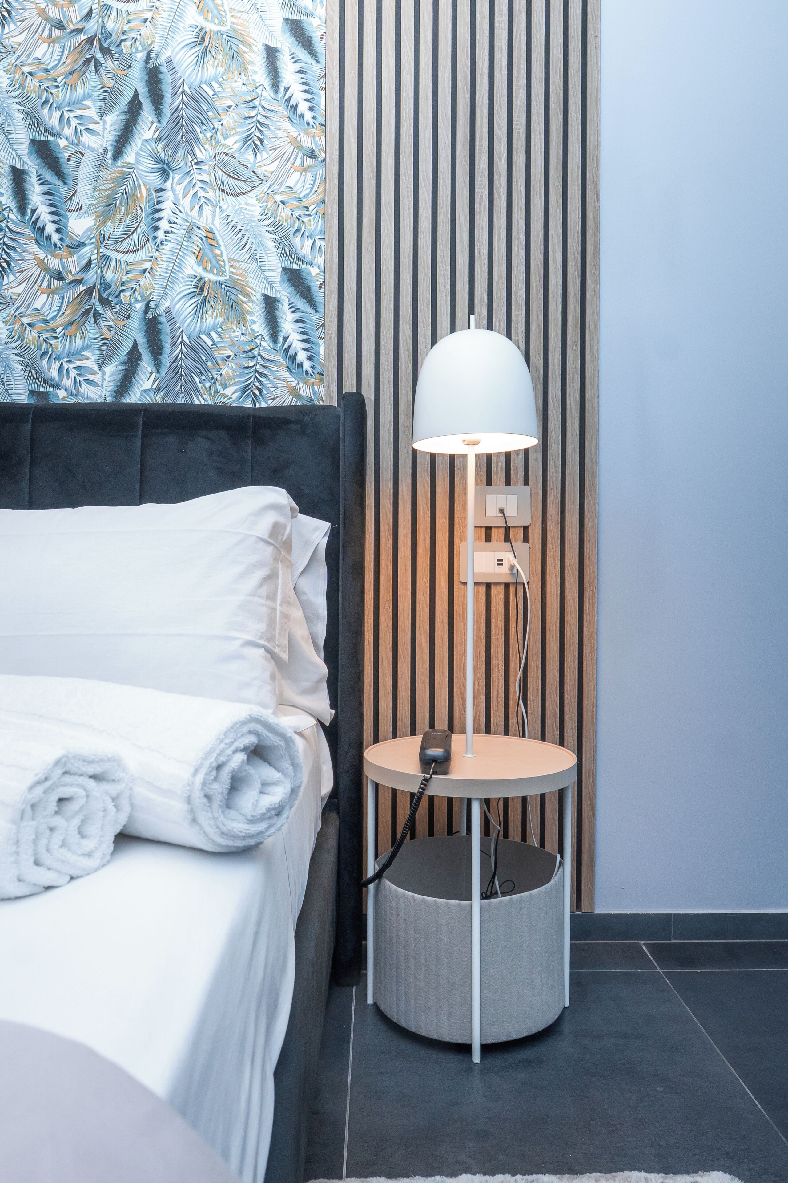 Una cama bien hecha con edredón blanco y toallas enrolladas, una lámpara moderna con pantalla blanca, y una pared con listones de madera verticales.