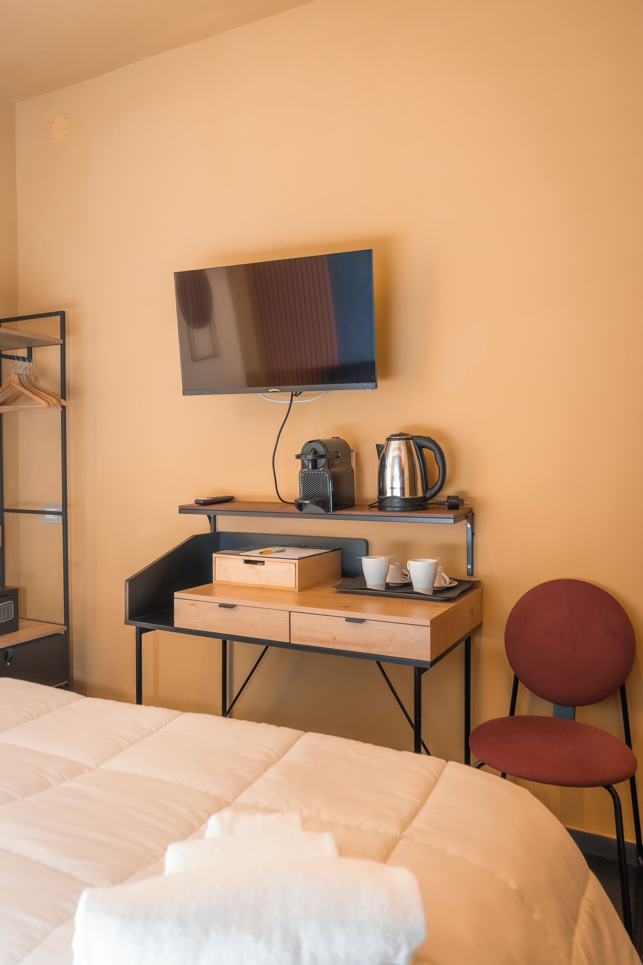 A hotel room with a bed, a desk with a kettle, cups, and a television on the wall.