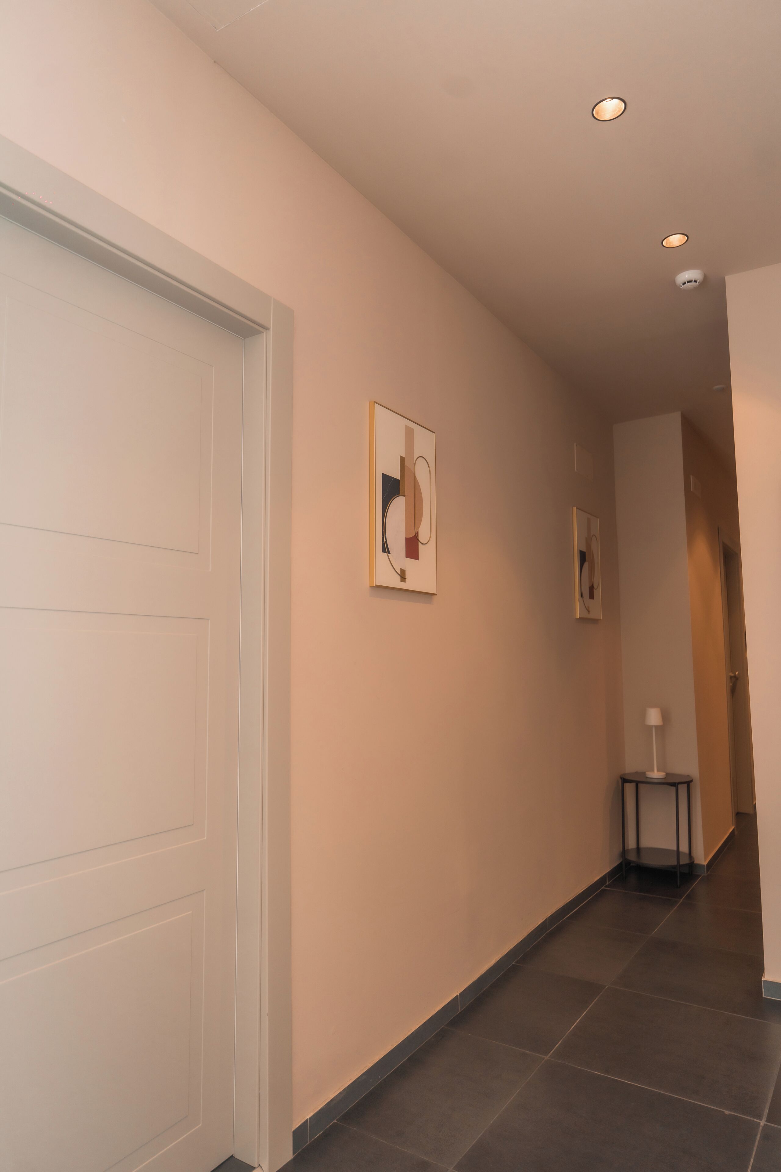A hallway with a white door, a small table with a lamp, and two framed artworks on the wall.