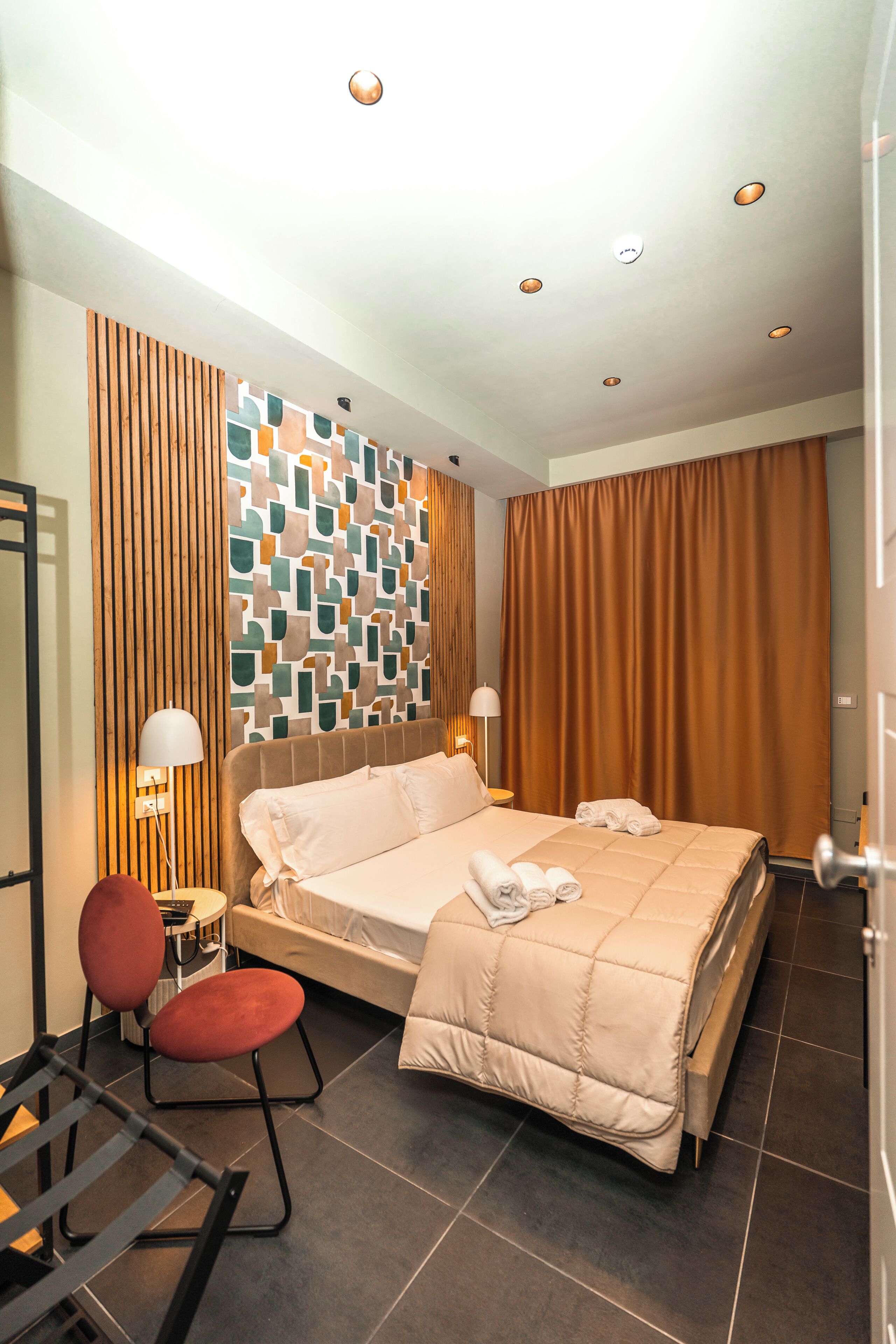 A hotel room with a bed, a desk, a chair, a lamp, and a wall with a geometric pattern.