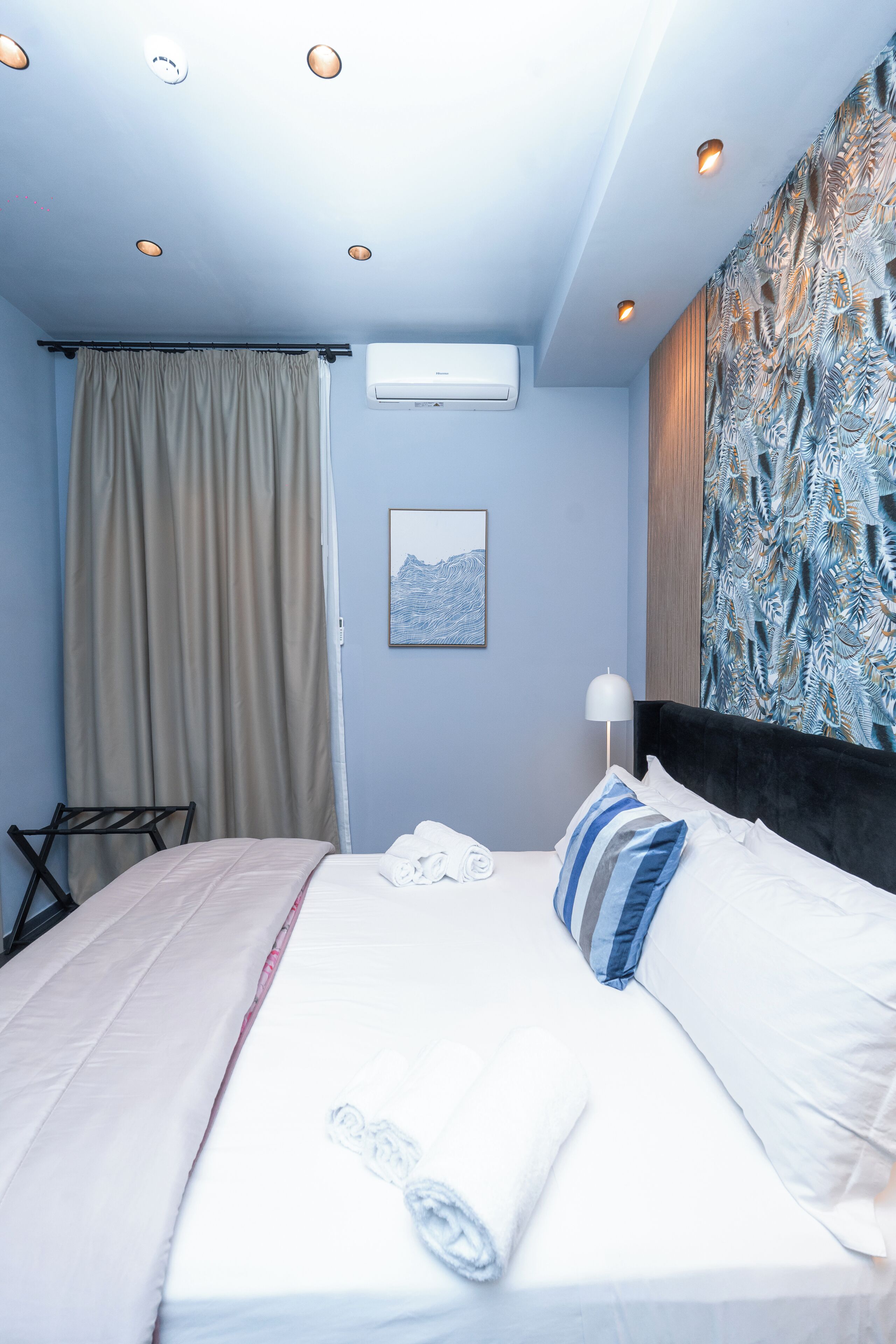 Una habitación de hotel con una cama, aire acondicionado montado en la pared, una ventana con cortinas, un cuadro enmarcado y un papel tapiz estampado.