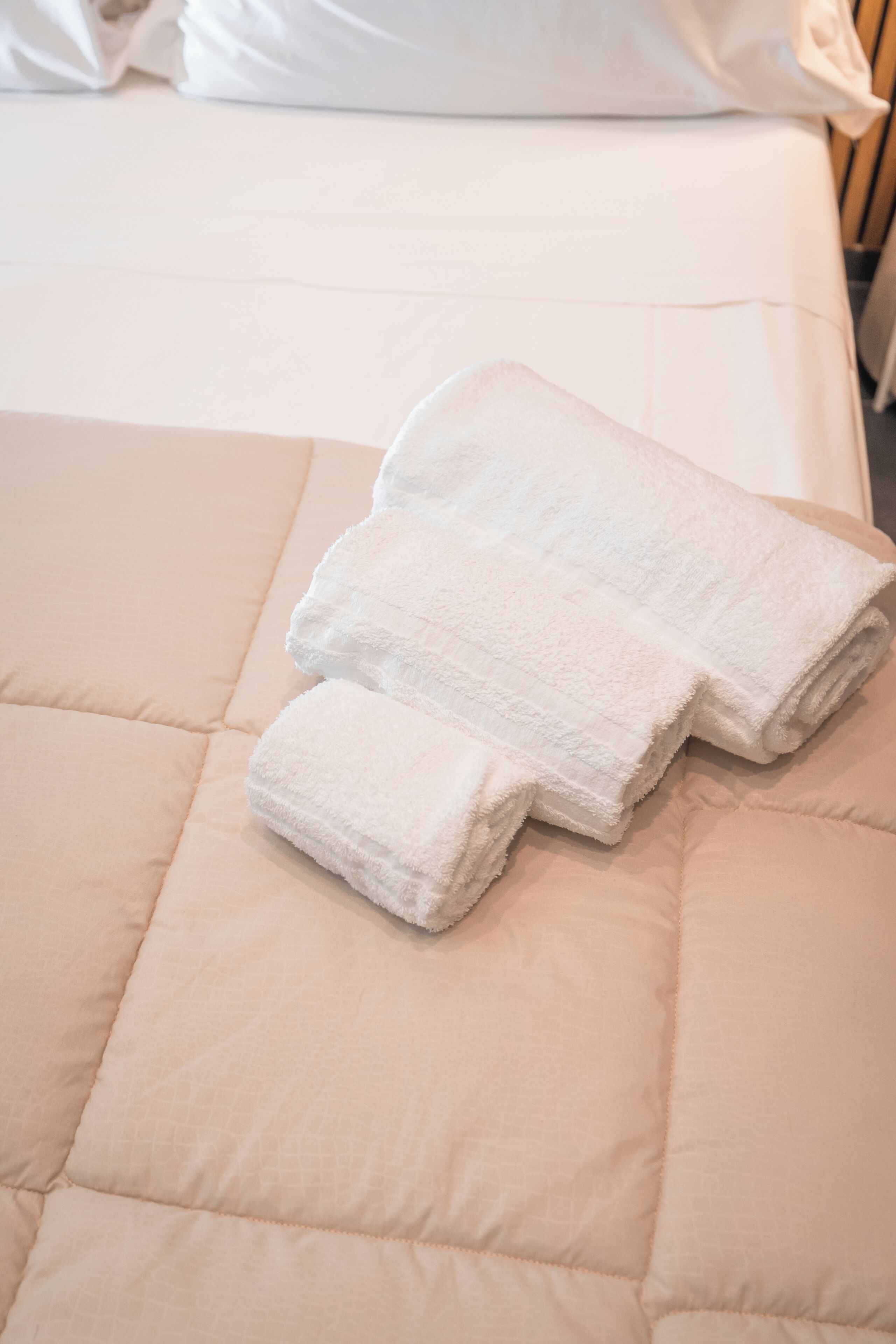 A bed with a beige quilted cover and three neatly folded white towels placed on it.