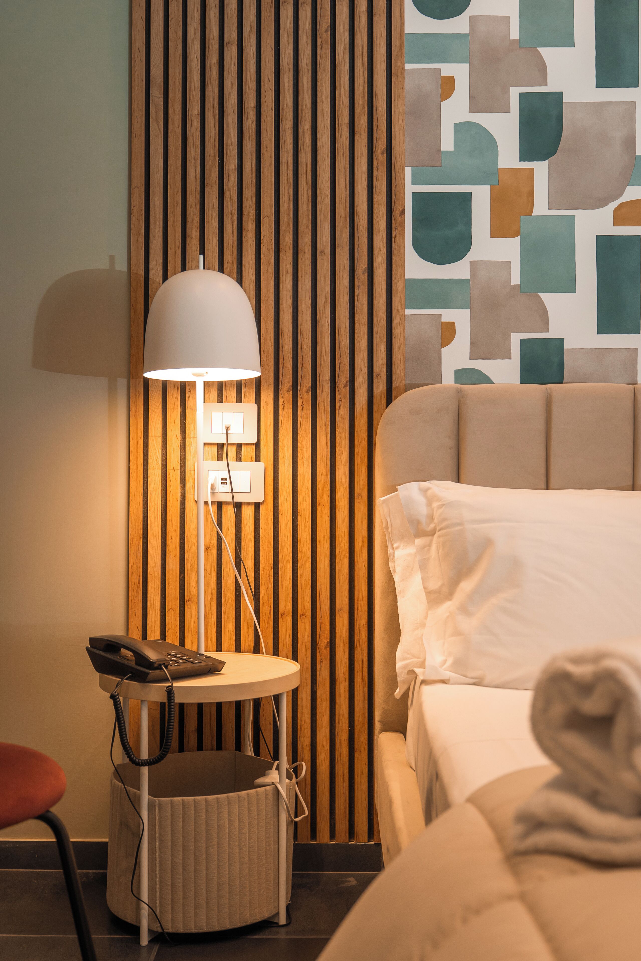 Une chambre d’hôtel comprenant un lit, une table de chevet avec une lampe, un téléphone et un mur orné de lattes de bois verticales et d’œuvres d’art murales abstraites.
