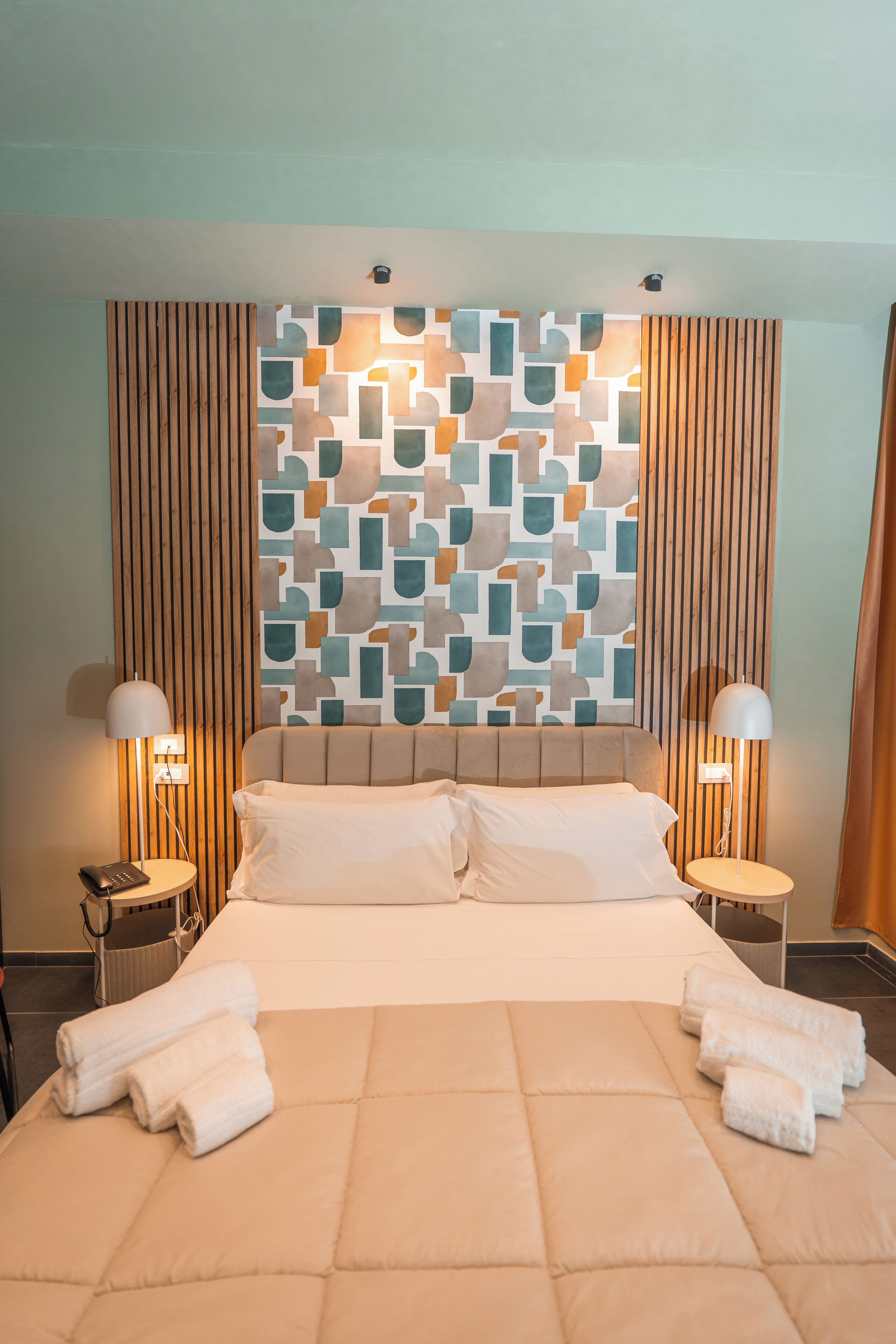 Habitación de hotel con una cama grande, dos lámparas de noche, una pared con diseños geométricos y una ventana con cortinas.