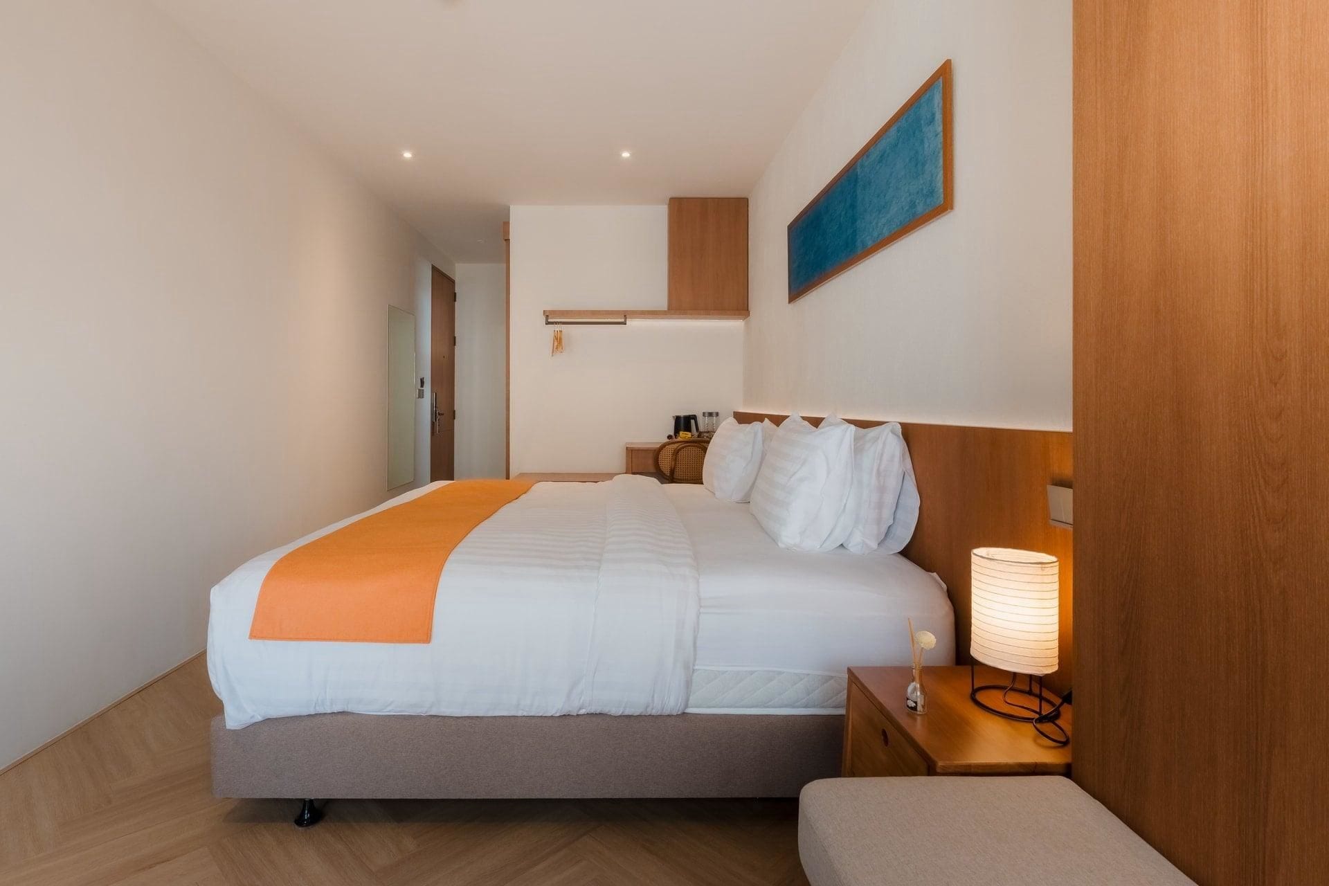 A hotel room with a bed, bedside table, lamp, and a painting on the wall.